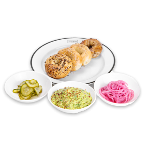 Bagels on a white plate with pickles, guacamole, and onions
